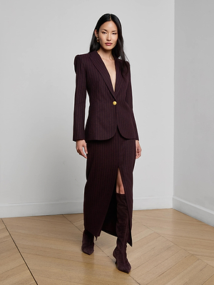 A woman stands indoors on a wooden floor, wearing the Sheila Longline Blazer with gold button detailing and a matching long skirt with a front slit. She completes the look with heeled boots and sleek, long hair.