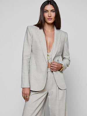 A woman with long brown hair poses confidently against a white background, wearing the Sheila Longline Blazer—an Italian wool-blend pinstripe suit with a deep neckline and gold accessories.