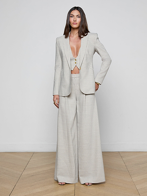 A woman stands against a white wall wearing the Sheila Longline Blazer, a light grey Italian wool-blend pinstripe suit jacket, paired with wide-leg trousers and neutral-toned heels. Her long straight hair falls over her shoulders on the wooden floor.