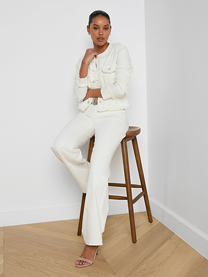 A woman poses confidently in the Yarin Collarless Fray Jacket, an all-white textured blazer. Seated on a wooden stool against a white wall, she rests one hand on her chin and the other on her lap, completing the look with sandals.