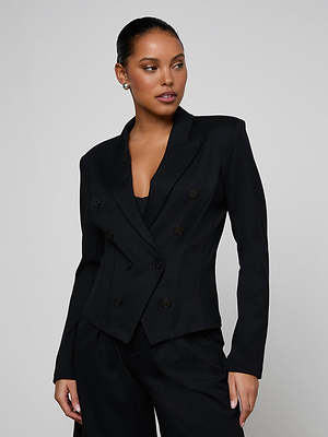 A woman with dark hair pulled back wears the Emerson Ponte Blazer—black, double-breasted, and sculpted—with matching pants. She stands against a plain gray background, looking to her left with a confident expression.