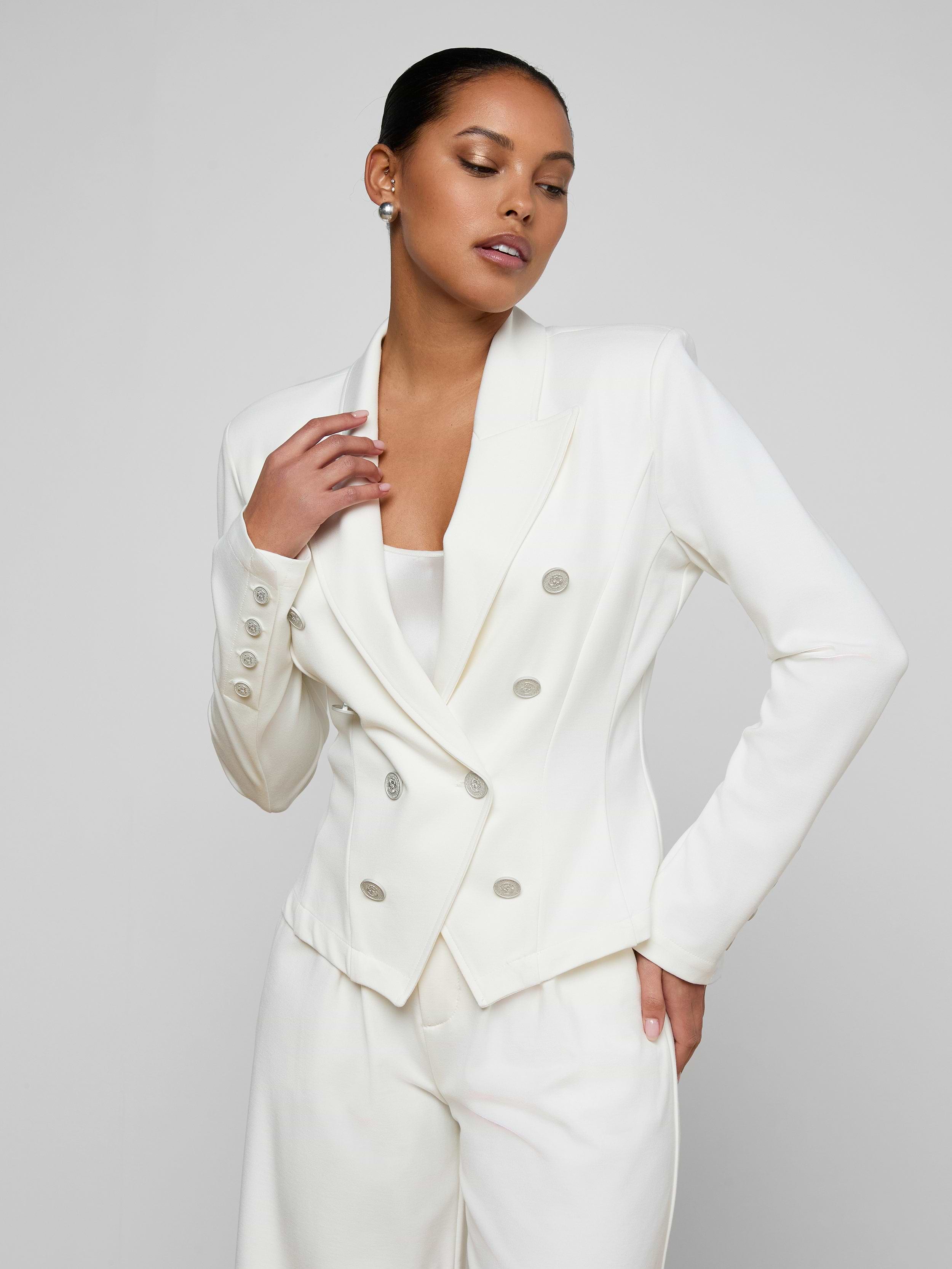 Wearing the Emerson Ponte Blazer in white with matching pants, a woman poses confidently against a plain, light background.