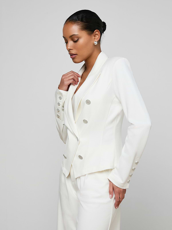 A woman models the Emerson Ponte Blazer—a tailored, white double-breasted design with silver buttons—against a plain, light background. Her hair is pulled back as she looks down, one hand in her pocket and the other gently touching the blazer.