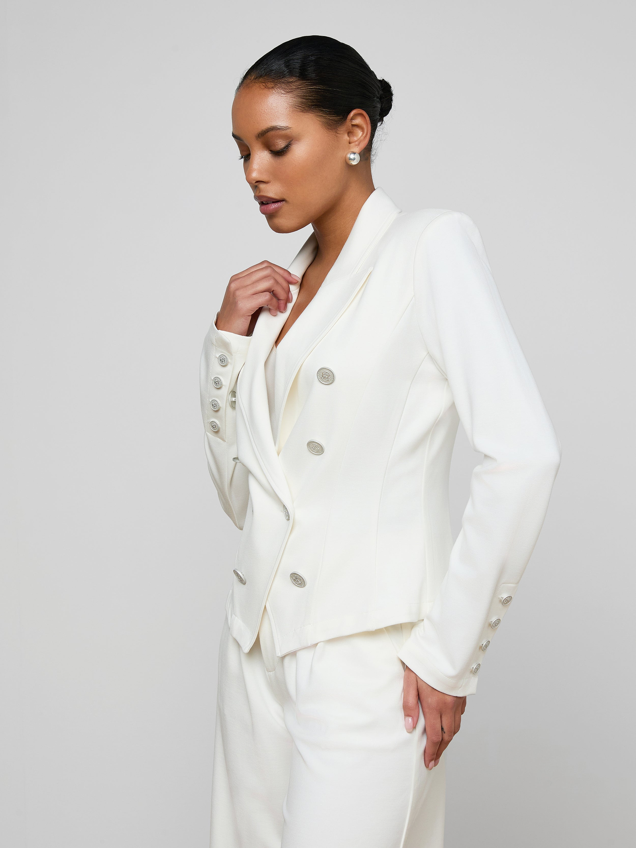 A woman models the Emerson Ponte Blazer—a tailored, white double-breasted design with silver buttons—against a plain, light background. Her hair is pulled back as she looks down, one hand in her pocket and the other gently touching the blazer.
