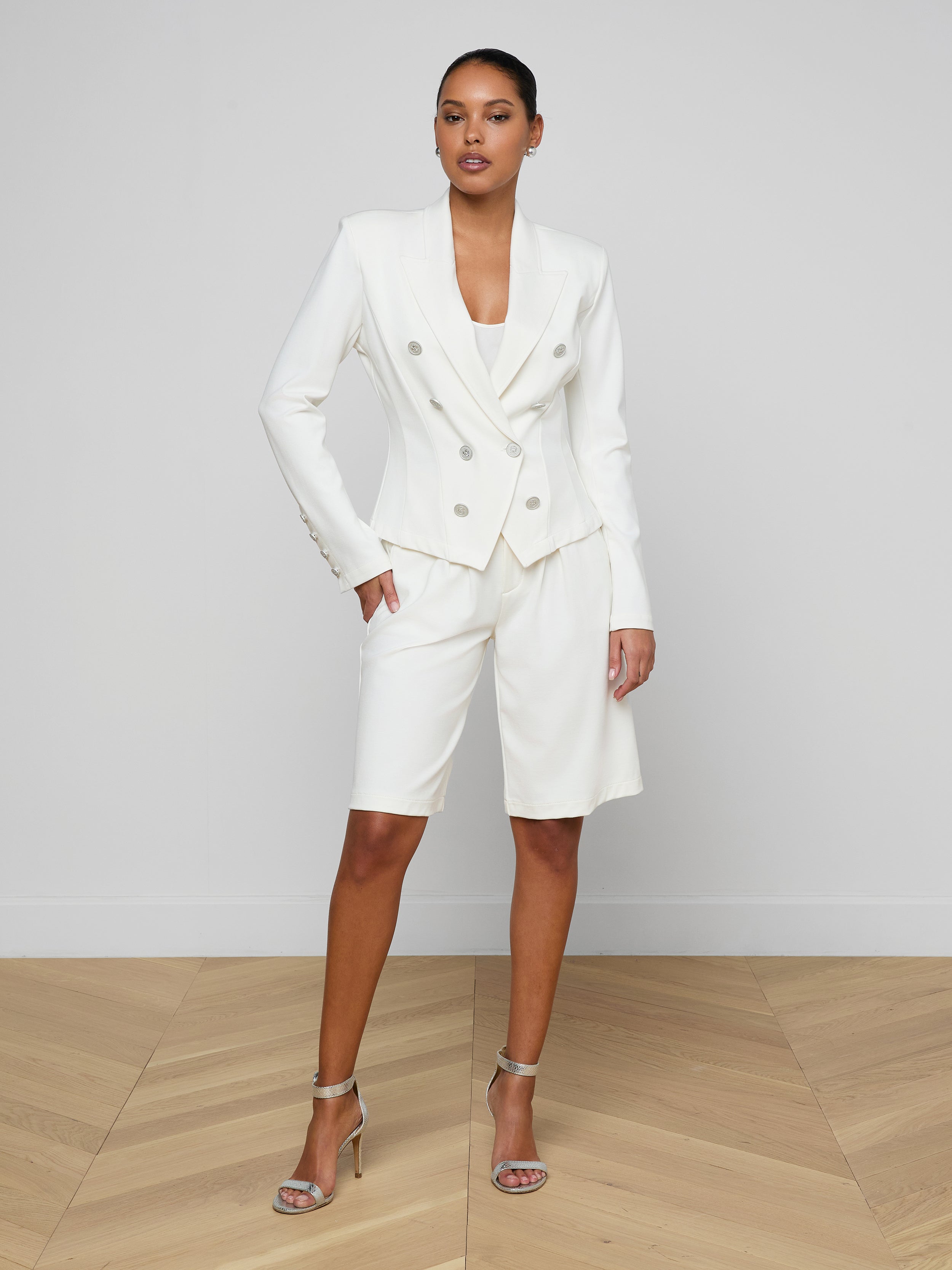 Styled in the Emerson Ponte Blazer—a tailored double-breasted design in white with silver buttons—this woman pairs it with matching knee-length super stretch ponte shorts and strappy high-heeled sandals, standing against a plain white background.