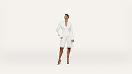 A woman stands confidently wearing a white double-breasted blazer, matching knee-length shorts, and high-heeled sandals against a plain white background.