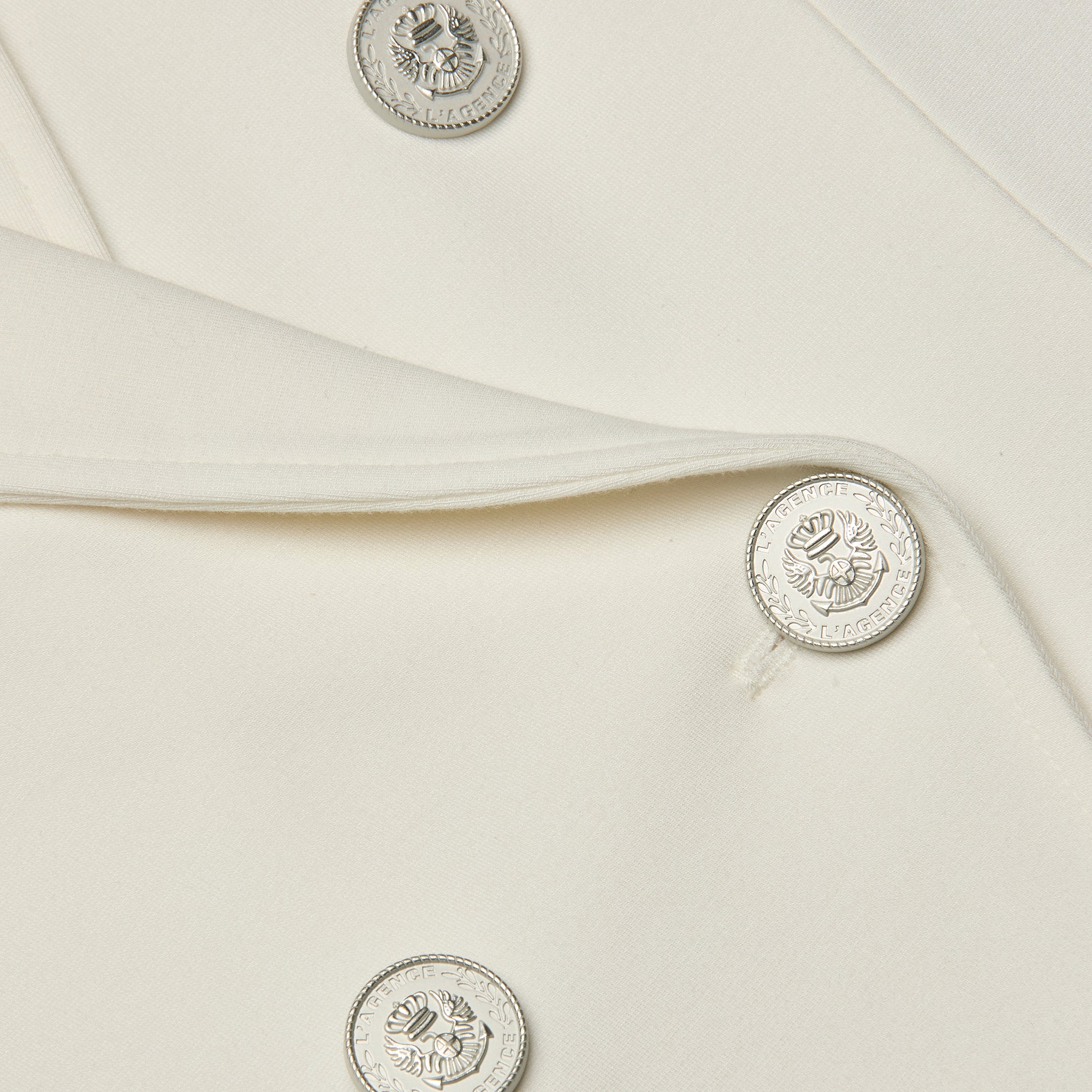 Close-up of the Emerson Ponte Blazer in white, showcasing shiny silver buttons with an embossed emblem. The smooth fabric and a portion of the lapel are visible.