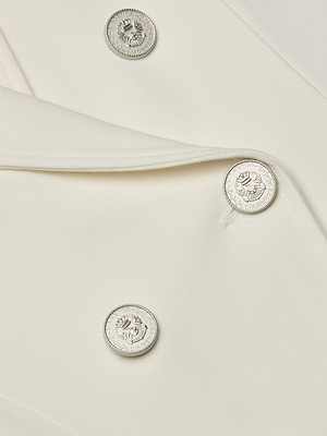 Close-up of the Emerson Structured Blazer in off-white, showcasing shiny silver lion-embossed buttons. Smooth fabric and precise stitching emphasize its sculpted, double-breasted silhouette.