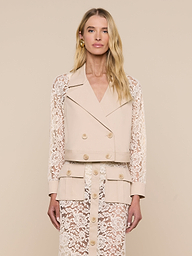 A woman with long blonde hair and a neutral expression stands facing forward against an off-white background, wearing the Novelle Lace Trench—a beige double-breasted blazer featuring floral lace sleeves—paired with a matching lace skirt.