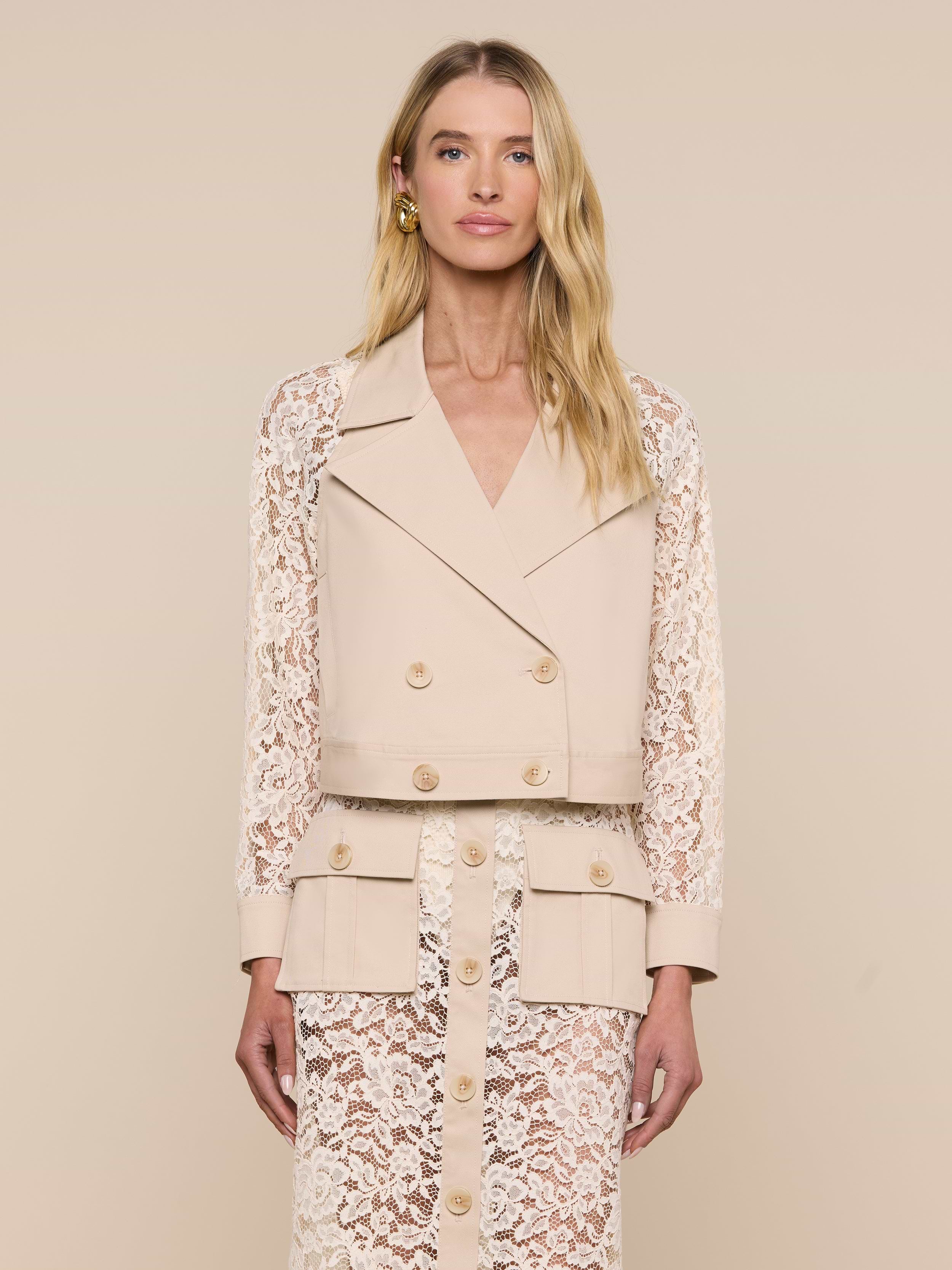 A woman with long blonde hair and a neutral expression stands facing forward against an off-white background, wearing the Novelle Lace Trench—a beige double-breasted blazer featuring floral lace sleeves—paired with a matching lace skirt.