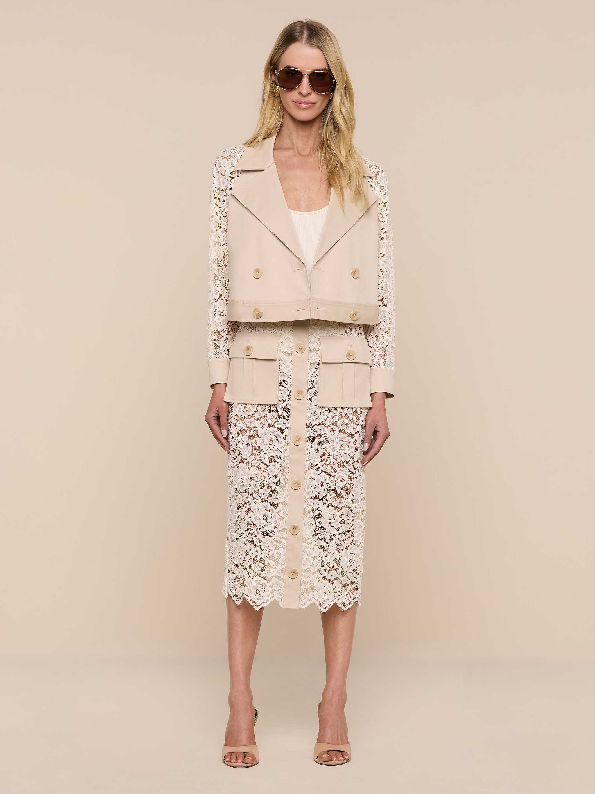 A woman wearing the Novelle Lace Trench, featuring floral lace sleeves, along with a matching beige top and a beige lace skirt with buttons, stands against a plain white background.