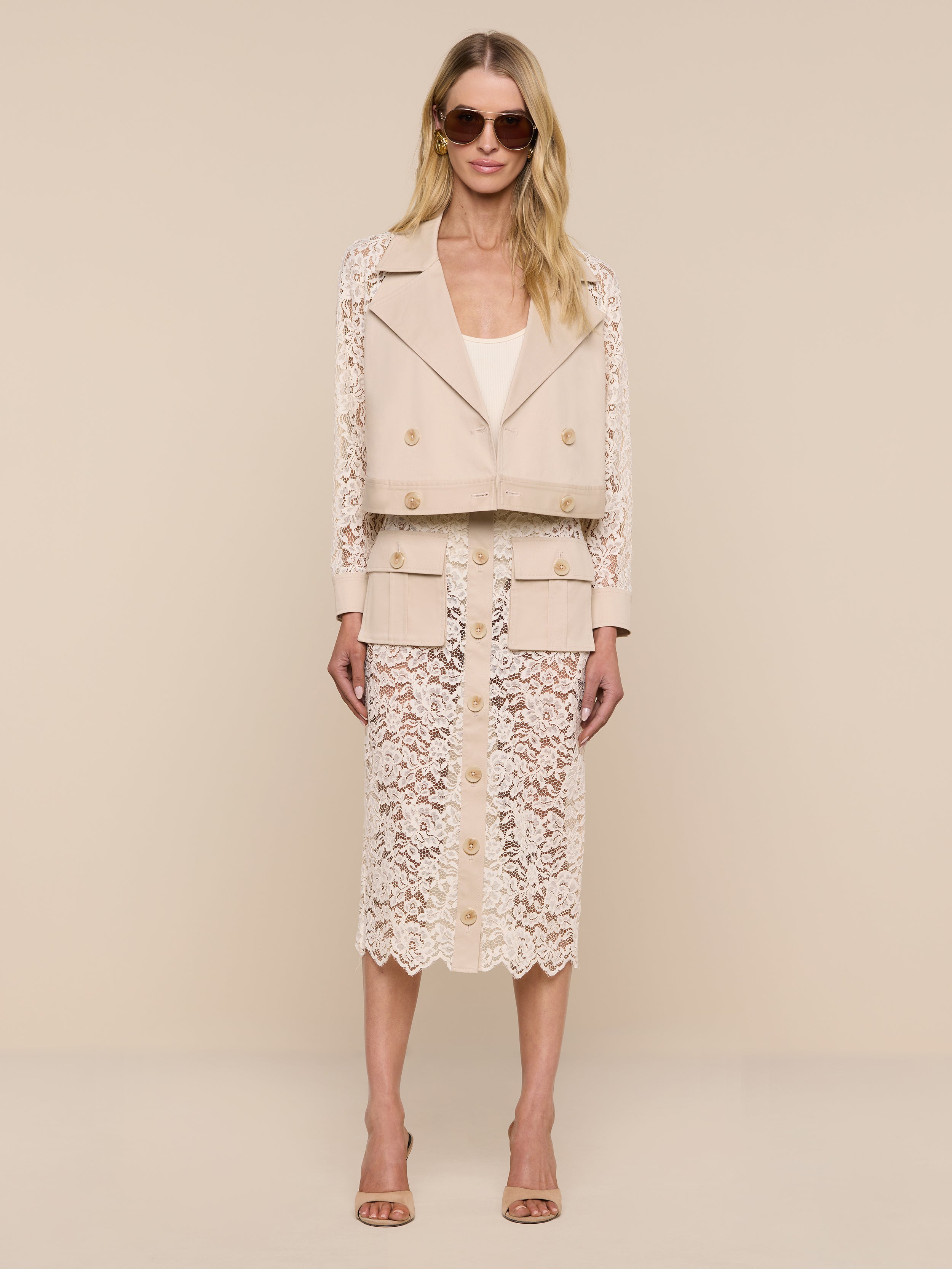 A woman wearing the Novelle Lace Trench, featuring floral lace sleeves, along with a matching beige top and a beige lace skirt with buttons, stands against a plain white background.