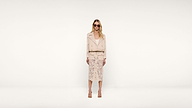 Woman wearing sunglasses, a light-colored jacket, and a lace skirt stands against a plain white background, facing the camera with a neutral expression.