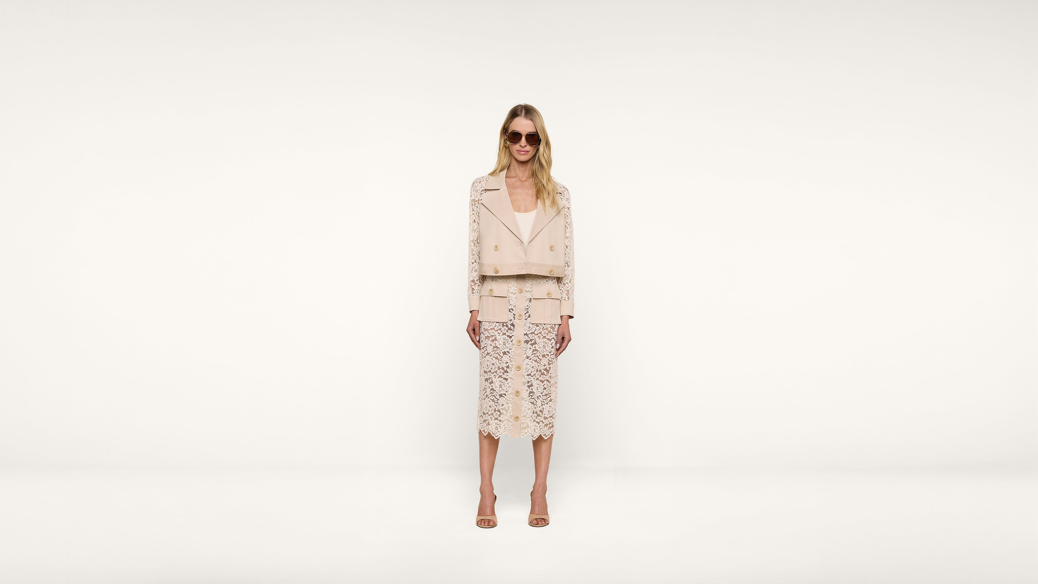 Woman wearing sunglasses, a light-colored jacket, and a lace skirt stands against a plain white background, facing the camera with a neutral expression.