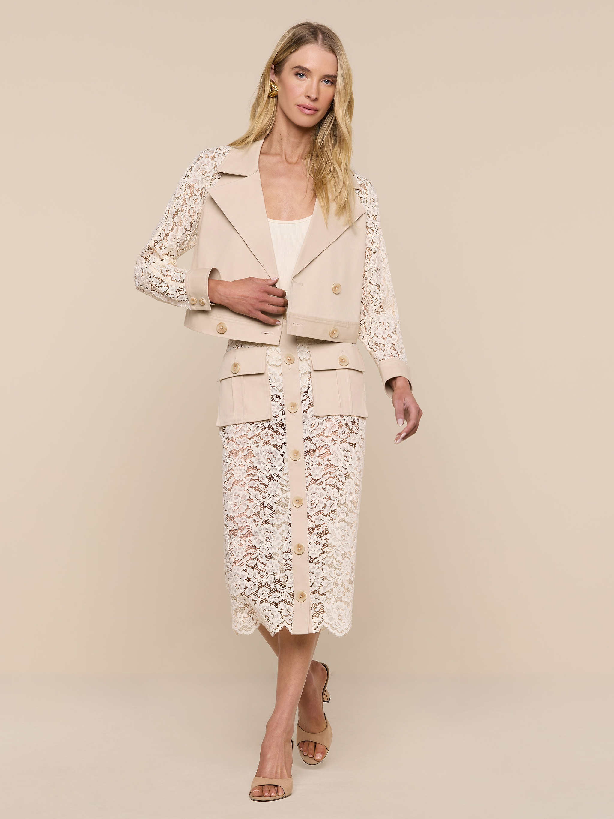 A woman stands against a plain white background, wearing the Novelle Lace Trench—a light beige, cream lace trench with floral lace sleeves. She completes her look with a matching top and skirt, open-toed sandals, and her long blonde hair worn down.