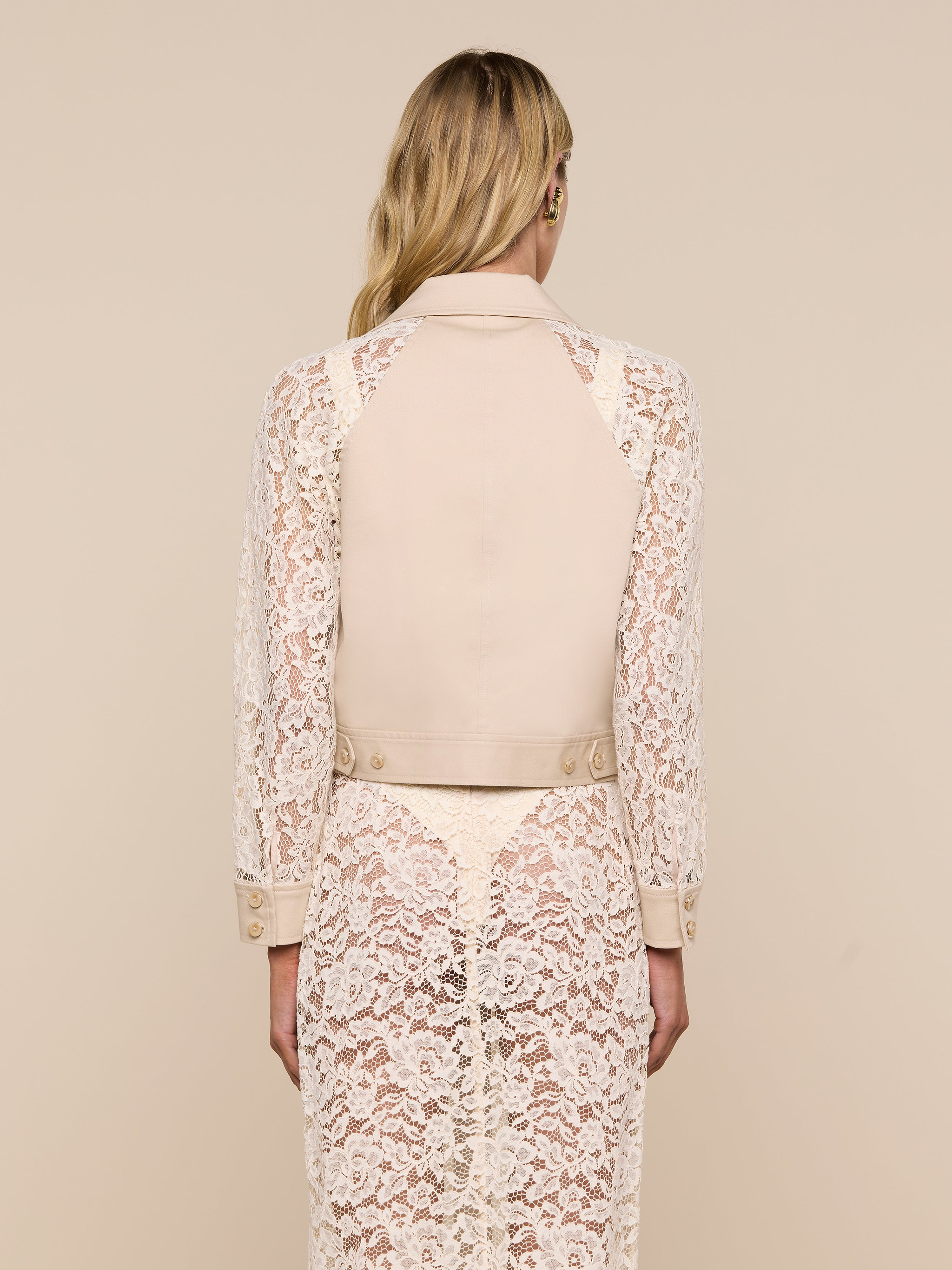 A woman with long blonde hair, seen from behind, wears the Novelle Lace Trench—a light beige coat featuring floral lace sleeves—over a cream lace dress against a plain, light background.