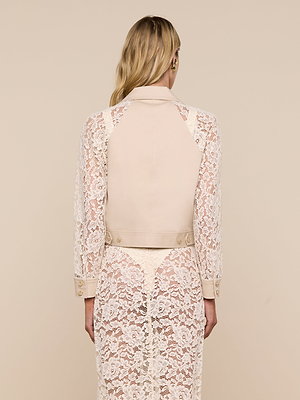 A woman faces away, wearing the Novelle Lace Trench—a beige trench coat with sheer white lace sleeves and a matching lace skirt. Her long, wavy blonde hair is down against a plain light beige background.