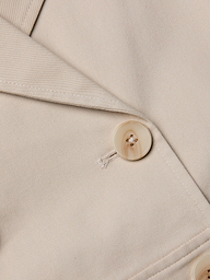 Close-up of the Novelle Lace Trench in beige, highlighting the lapel and a single light-colored button sewn with matching thread. The smooth-textured fabric and visible stitching details make this lightweight trench an ideal layer for refined looks.
