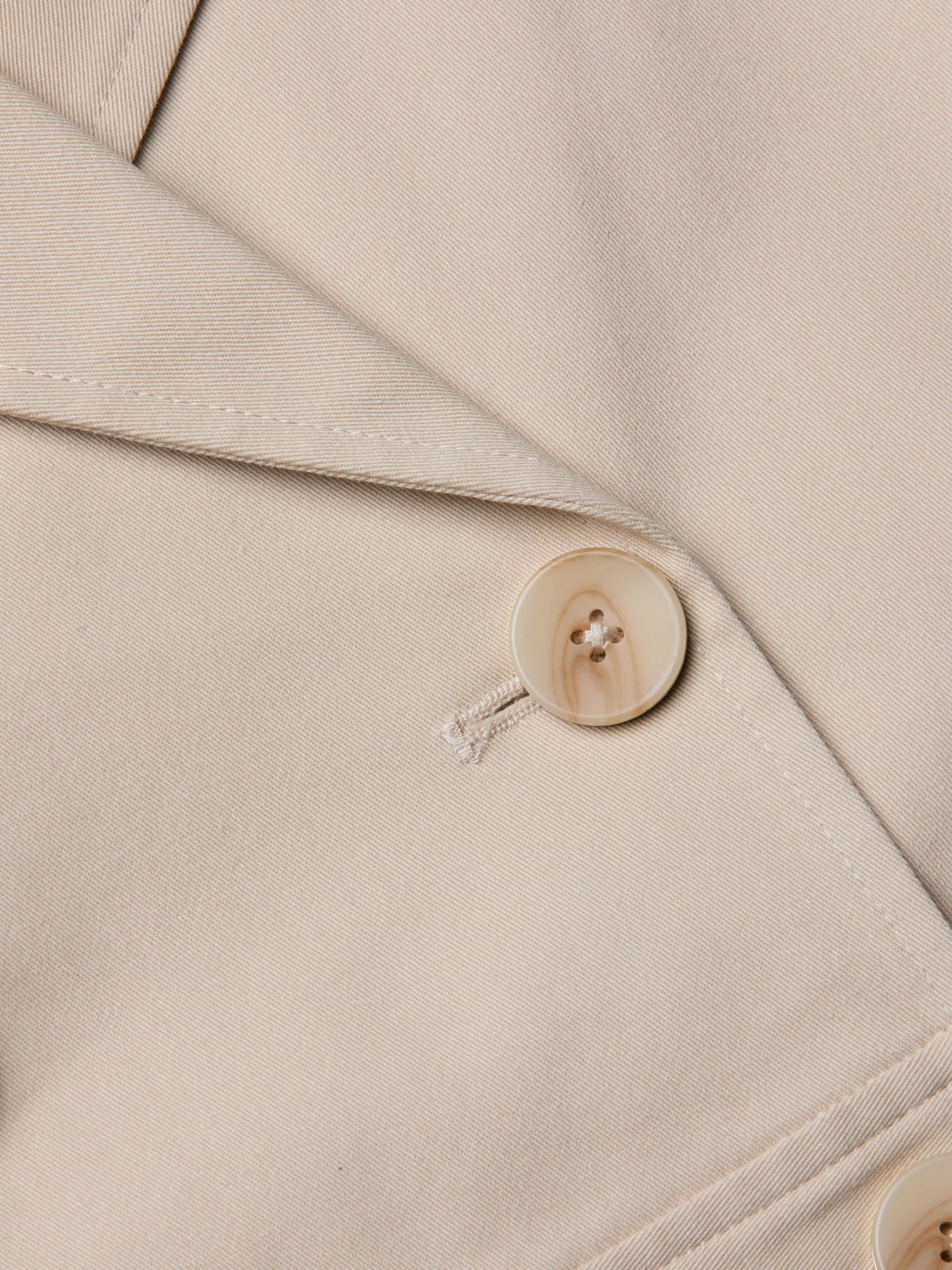 Close-up of the Novelle Lace Trench in beige, highlighting the lapel and a single light-colored button sewn with matching thread. The smooth-textured fabric and visible stitching details make this lightweight trench an ideal layer for refined looks.