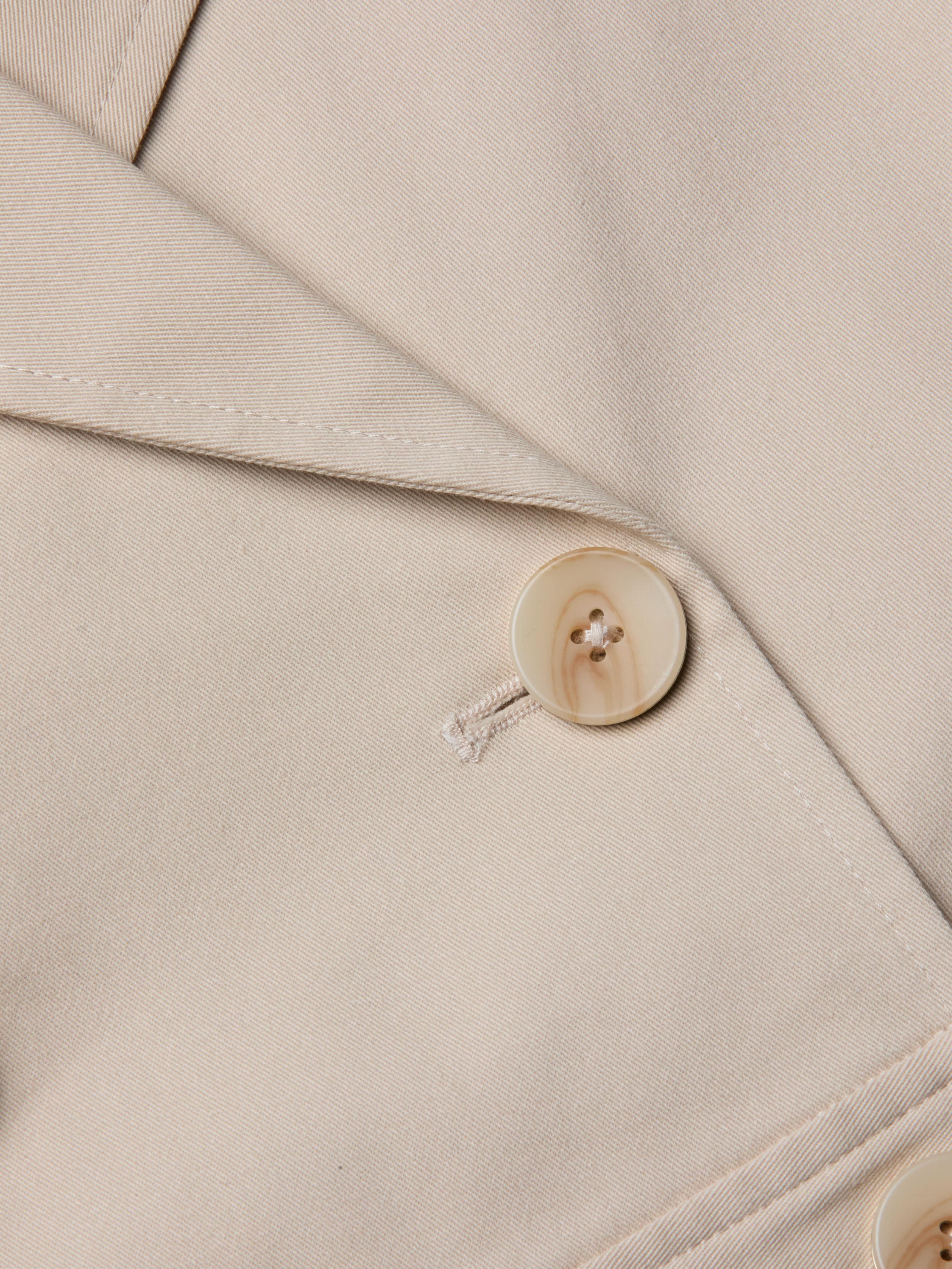 Close-up of the Novelle Lace Trench in beige, highlighting the lapel and a single light-colored button sewn with matching thread. The smooth-textured fabric and visible stitching details make this lightweight trench an ideal layer for refined looks.