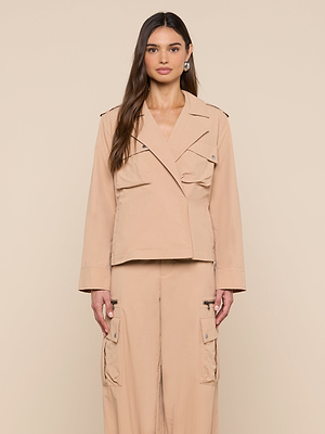 A woman with long brown hair wears the Calida Windbreaker Trench in beige, paired with matching cargo pants, standing against a plain beige background and facing the camera with a neutral expression.