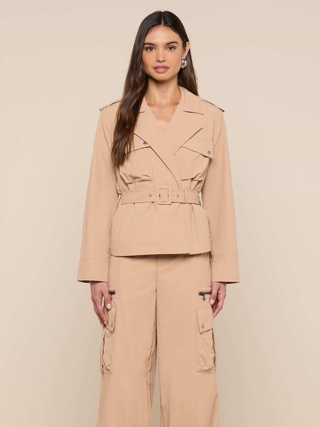 A woman stands facing forward against a plain, light background, wearing the Calida Windbreaker Trench—a beige, water-repellent jacket with a belted trench coat style—paired with matching cargo pants.