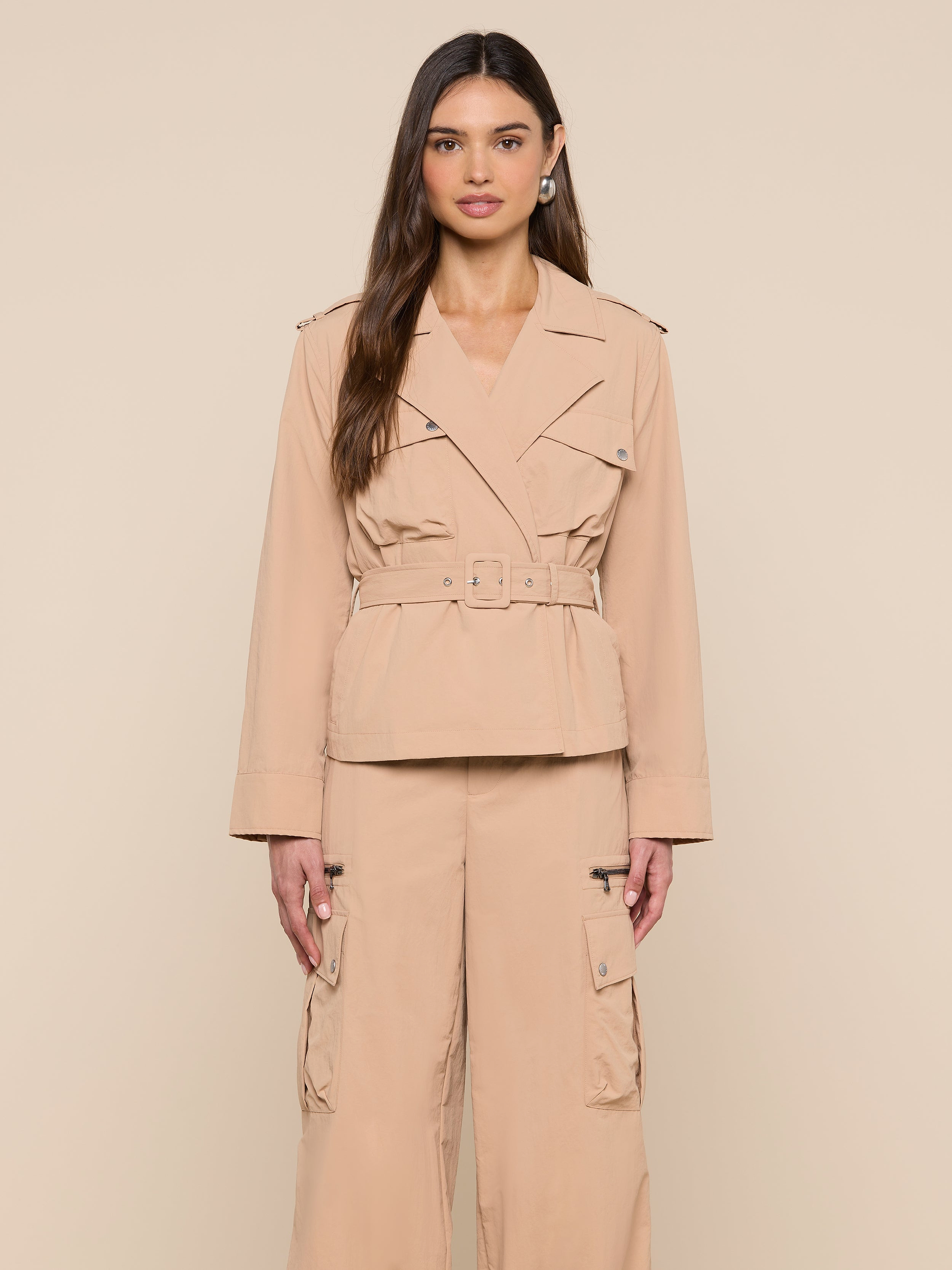 A woman stands facing forward against a plain, light background, wearing the Calida Windbreaker Trench—a beige, water-repellent jacket with a belted trench coat style—paired with matching cargo pants.