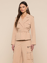 A woman stands against a plain light background, wearing the Calida Windbreaker Trench—a belted, beige utility jacket crafted from water-repellent nylon—paired with matching cargo pants. Her hair is down and her hands rest by her sides.