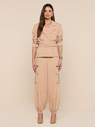 A woman stands in front of a white background, wearing the Calida Windbreaker Trench in beige, styled with matching cargo pants and tan heels. Her long hair is worn down, and she faces forward with a neutral expression.