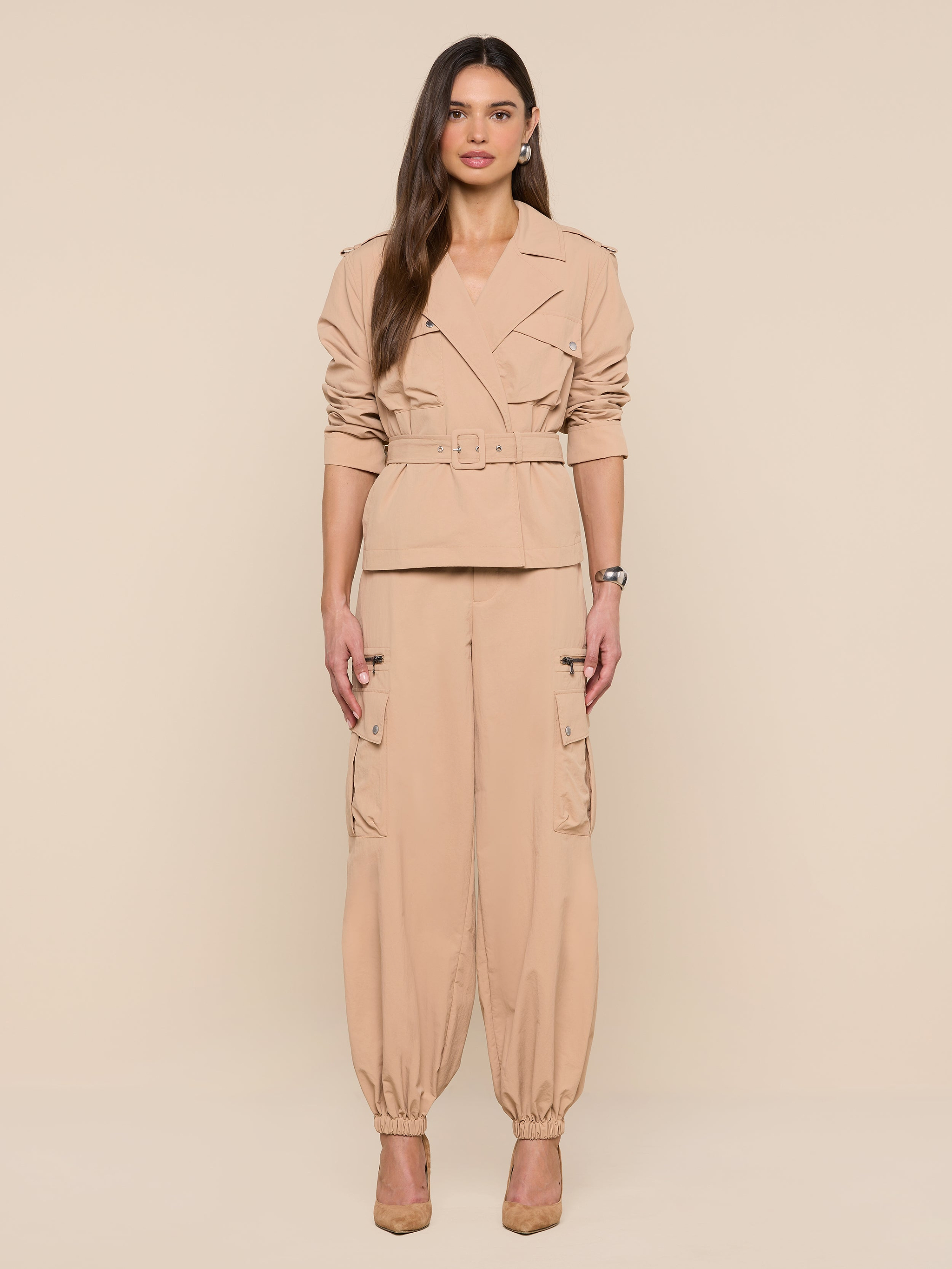 A woman stands in front of a white background, wearing the Calida Windbreaker Trench in beige, styled with matching cargo pants and tan heels. Her long hair is worn down, and she faces forward with a neutral expression.