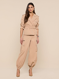 A woman with long brown hair strikes a relaxed, confident pose against a plain white background, wearing the Calida Windbreaker Trench in tan, paired with matching cargo pants and tan heels.
