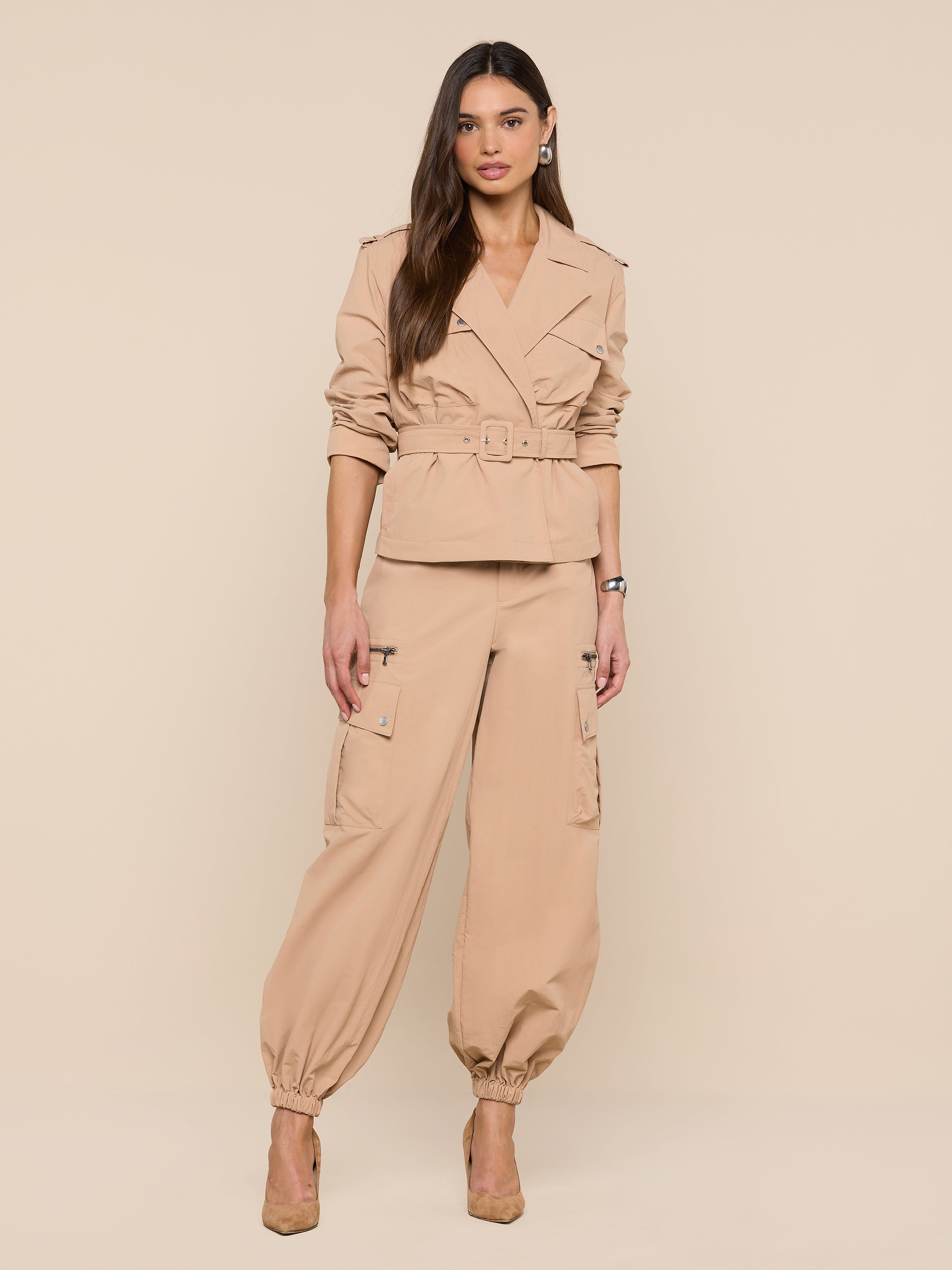 A woman with long brown hair strikes a relaxed, confident pose against a plain white background, wearing the Calida Windbreaker Trench in tan, paired with matching cargo pants and tan heels.