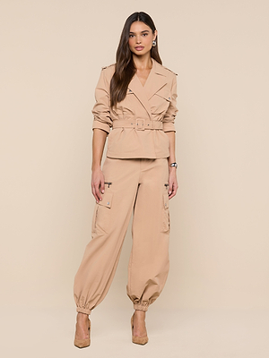 A woman poses against a beige backdrop wearing the Calida Windbreaker Trench and matching cargo pants. With long brown hair, beige heels, and minimal jewelry, she embodies effortless modern trench coat style.