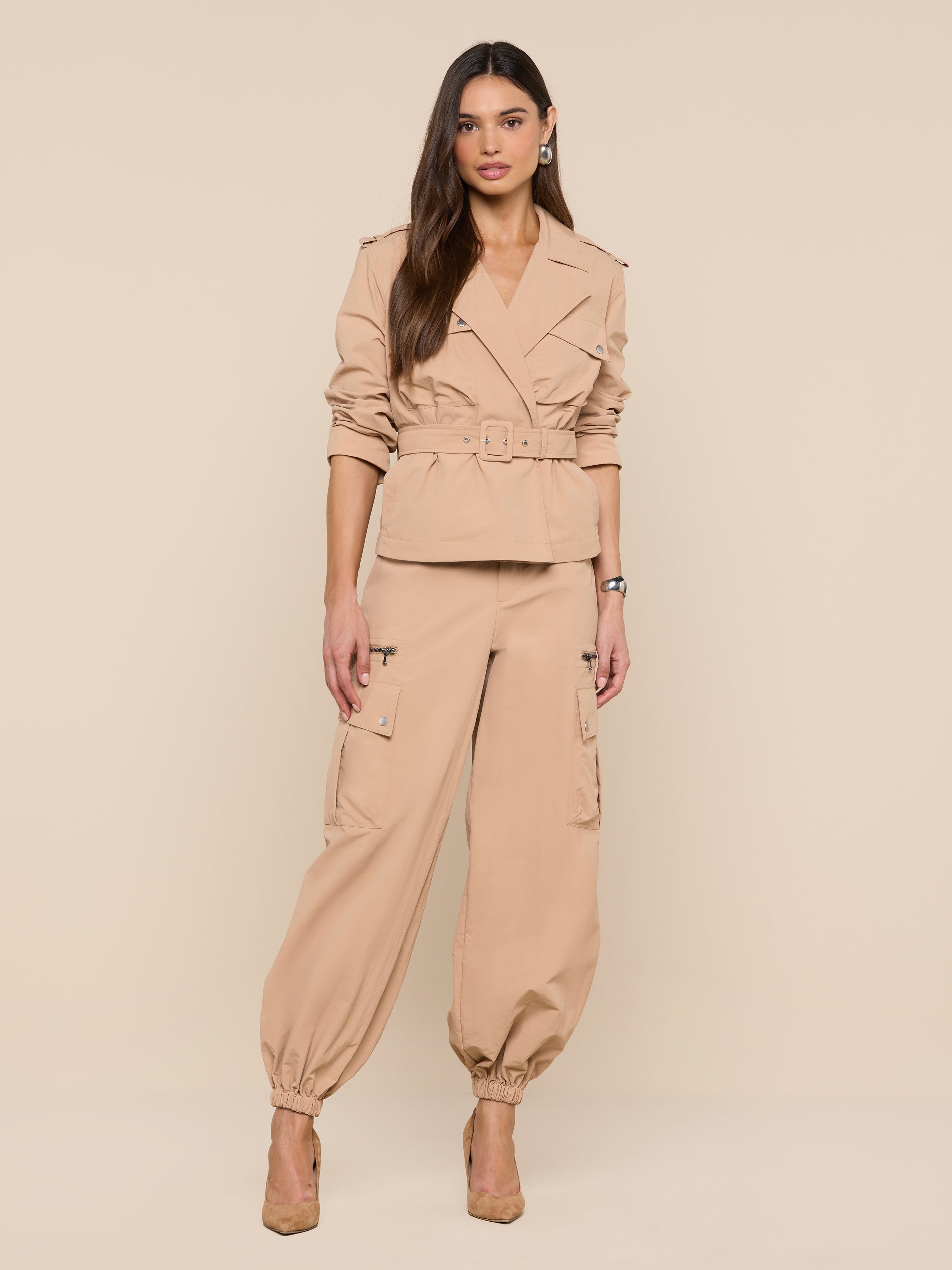 A woman with long brown hair strikes a relaxed, confident pose against a plain white background, wearing the Calida Windbreaker Trench in tan, paired with matching cargo pants and tan heels.