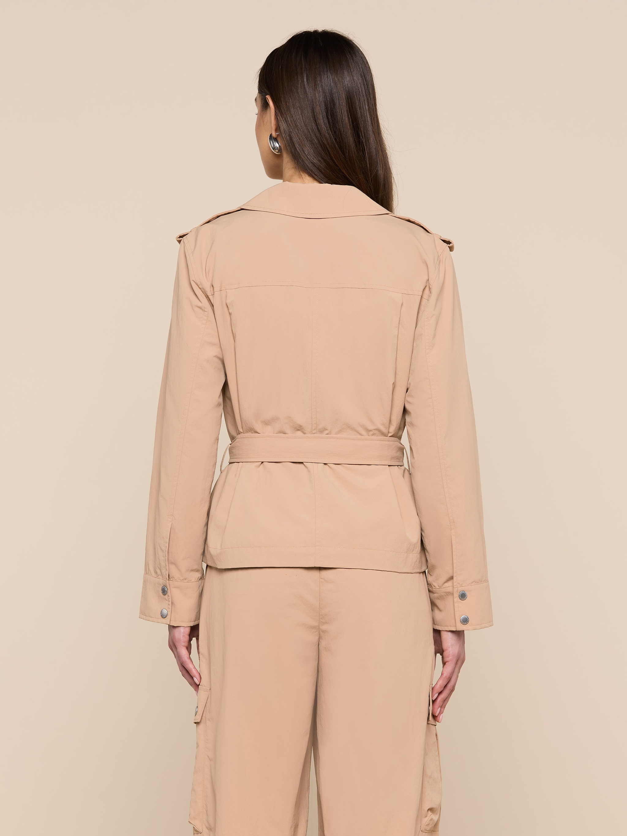 A person with long brown hair is standing facing away, wearing the Calida Windbreaker Trench—a light brown water-repellent trench coat with a belt—paired with matching pants. The background is plain and light-colored.