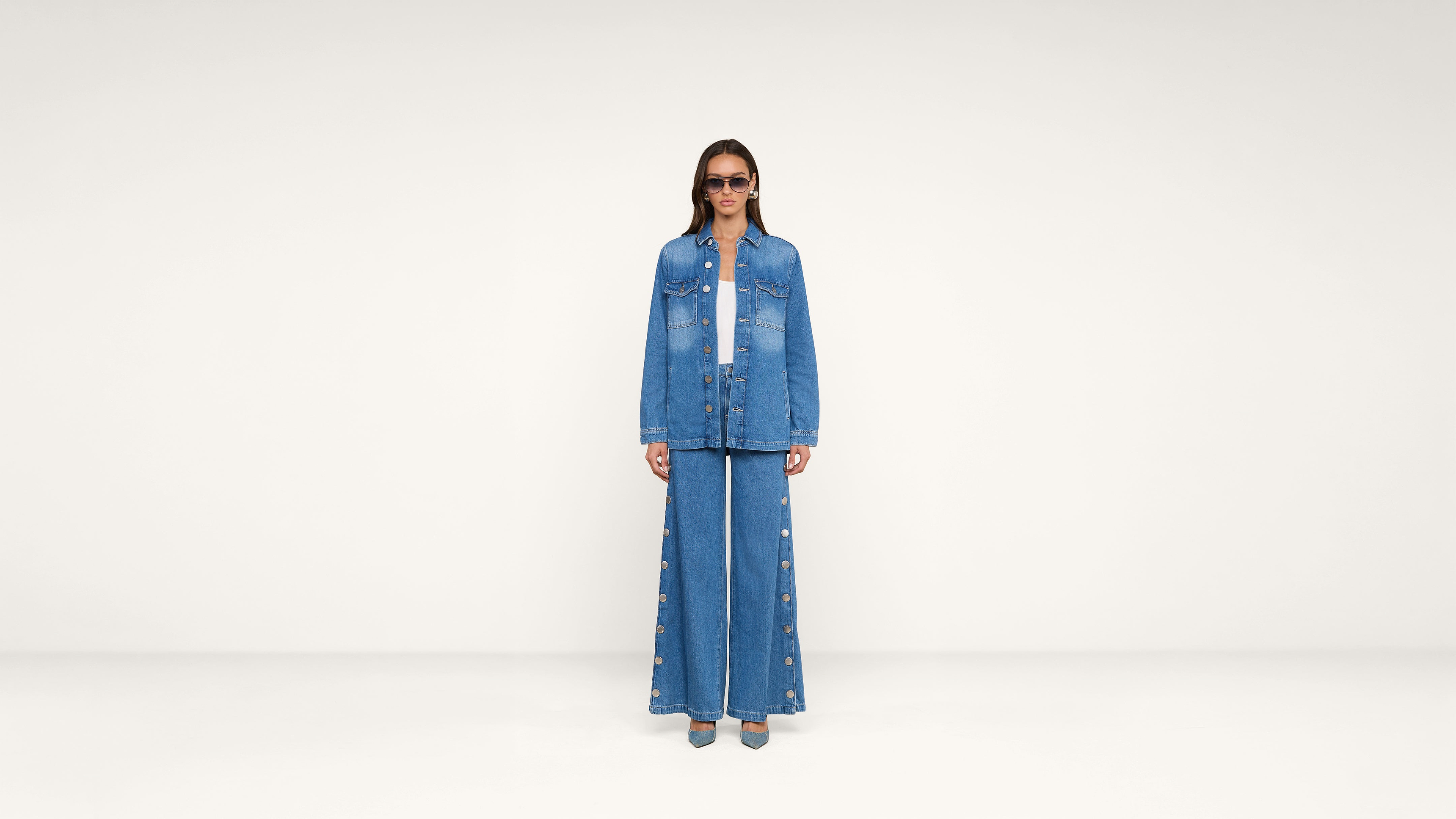 A person stands in a white room wearing blue sunglasses, a blue denim jacket, and wide-leg denim pants with buttons down the sides, along with blue shoes.