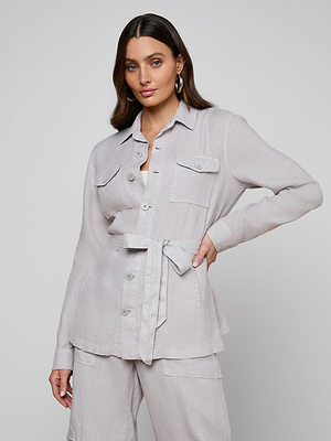 A woman with long brown hair poses in the Samaya Linen Jacket, a light beige utility style with a tie belt at the waist, paired with matching pants. She wears hoop earrings and stands against a plain, light gray background.
