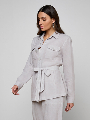 A woman with long brown hair wears the Samaya Linen Jacket—a light beige utility jacket with a tie belt, buttons, and front pockets—paired with matching pants, standing against a plain light gray background and looking down to the side.