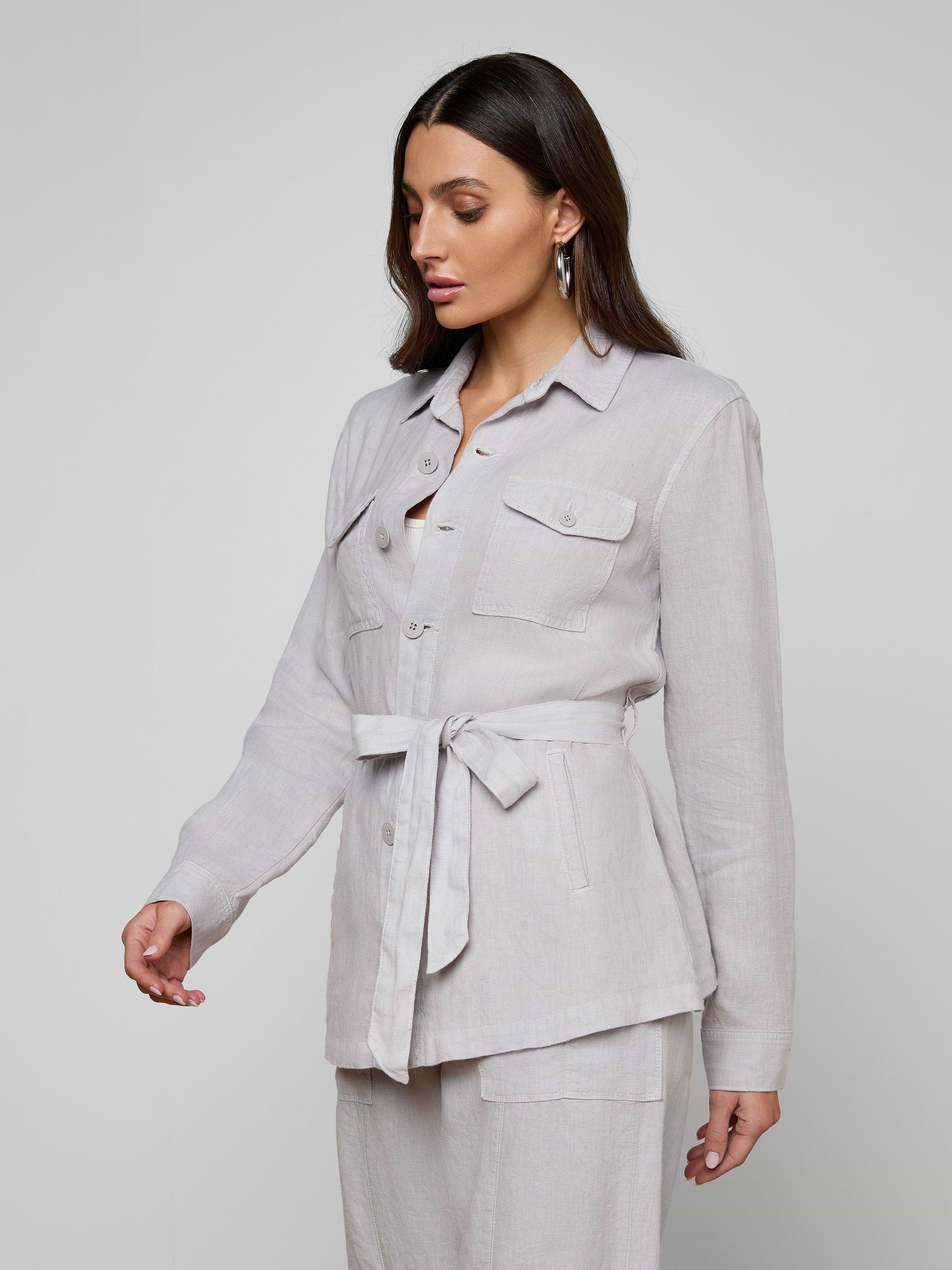 Wearing the Samaya Linen Jacket—a light-colored, versatile linen design with patch pockets—she pairs it with matching pants. Standing against a plain, off-white background, she looks down, her long dark hair cascading over her shoulders.