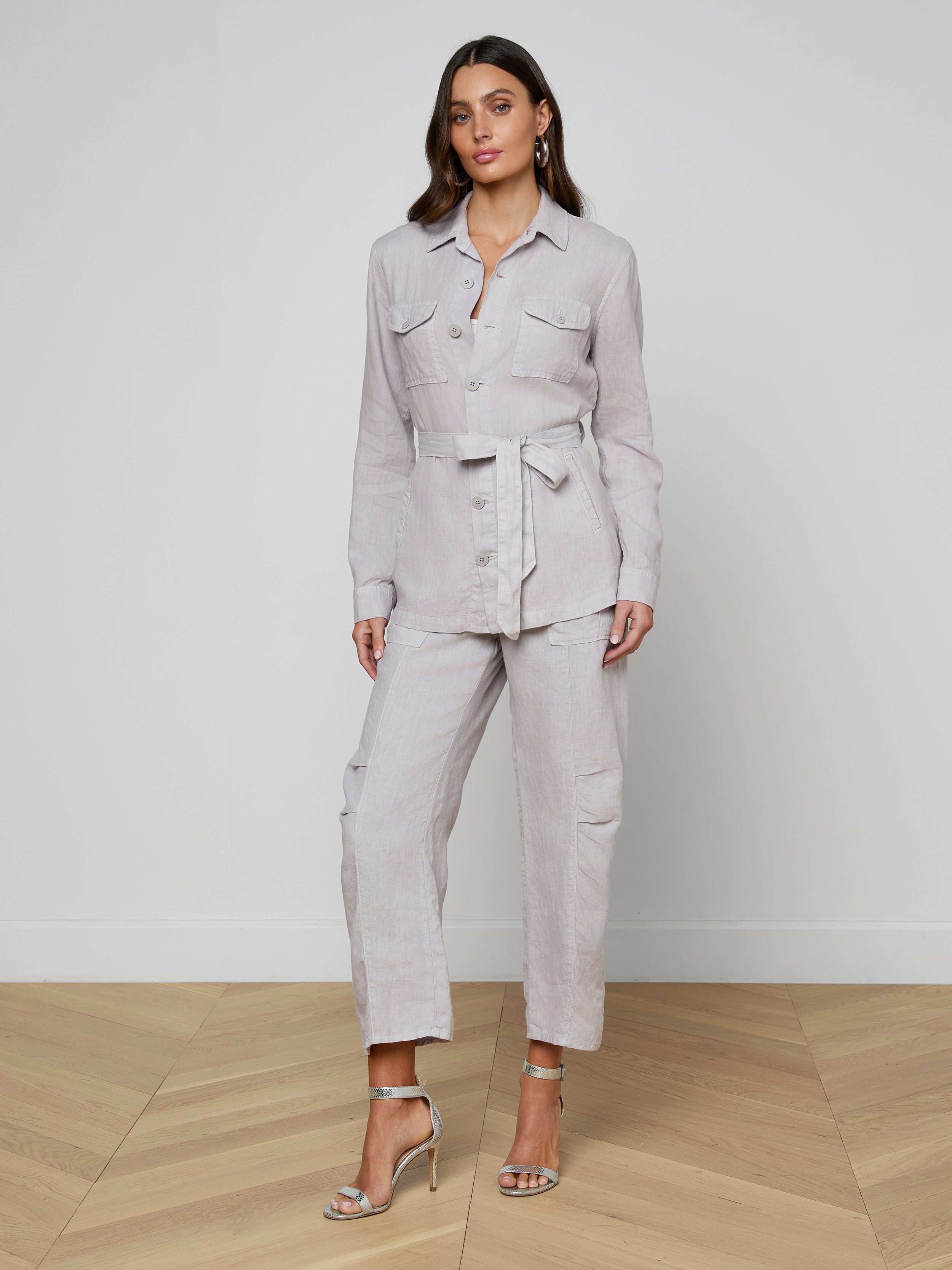 A woman stands facing forward against a plain white background, wearing the Samaya Linen Jacket in light gray, styled as a belted utility jumpsuit and paired with beige high-heeled sandals.