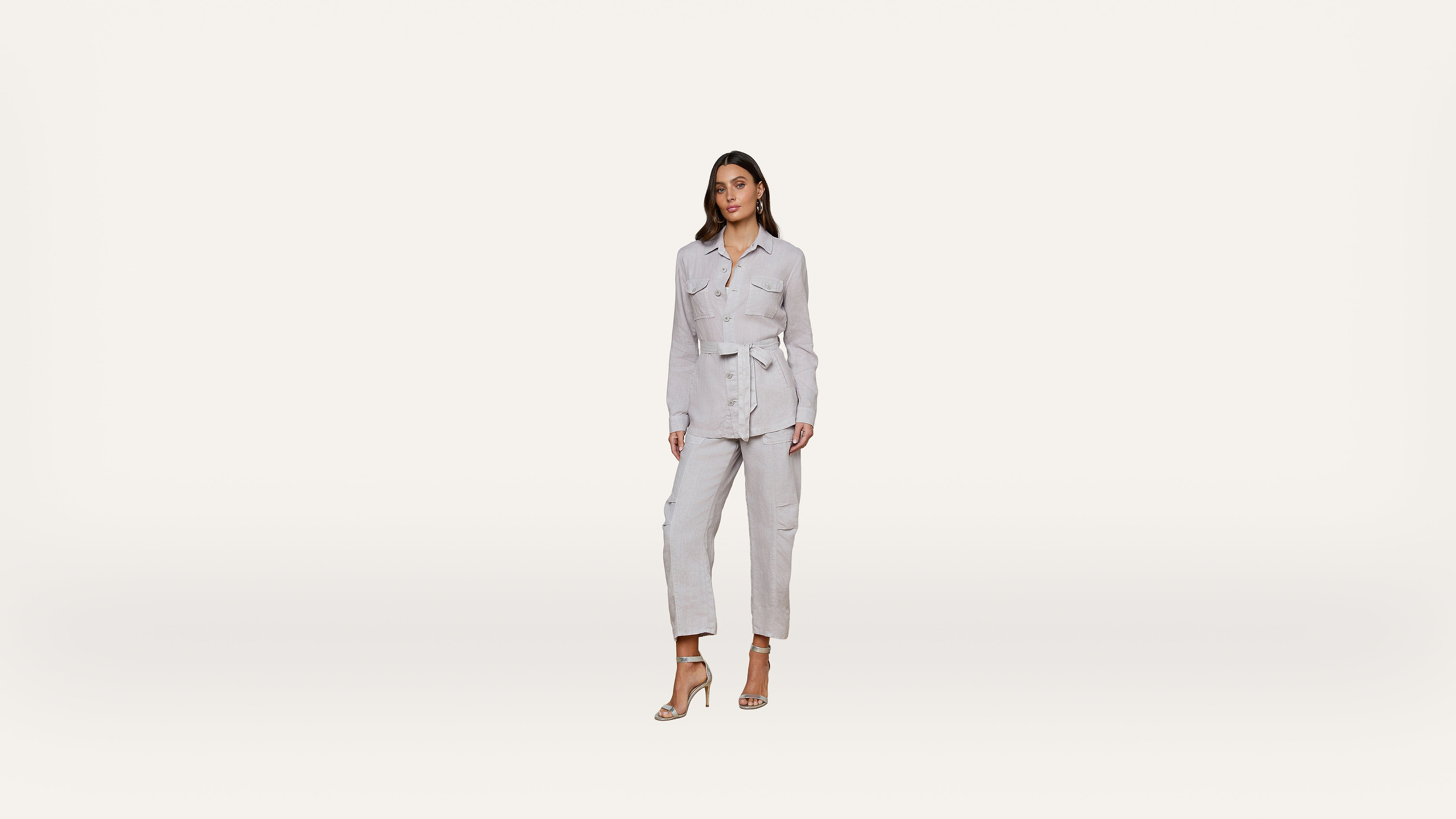 A woman wearing a light gray belted jacket and matching pants stands against a plain white background, posing in high-heeled sandals.