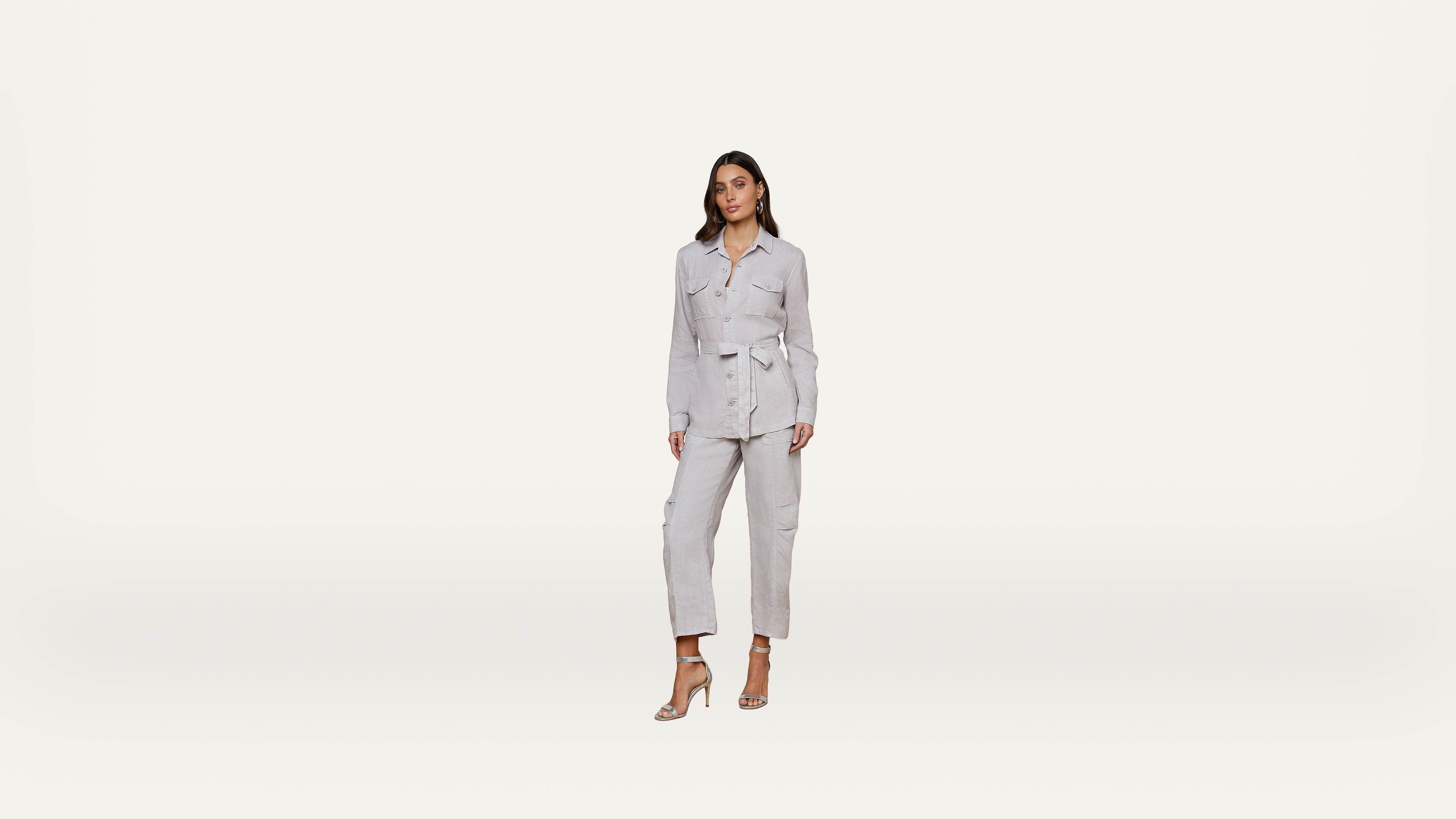 A woman wearing a light gray belted jacket and matching pants stands against a plain white background, posing in high-heeled sandals.