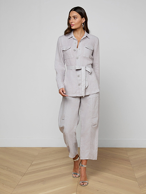 A woman stands indoors against a plain background, looking to the side while wearing the Samaya Linen Jacket in light gray with chest pockets and a tie belt, paired with matching loose pants and silver high-heeled sandals.