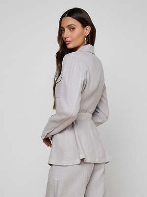 A woman with long brown hair wears silver hoop earrings and the Samaya Linen Jacket, a light gray style with a tie belt, standing slightly turned and looking over her shoulder against a plain white background.