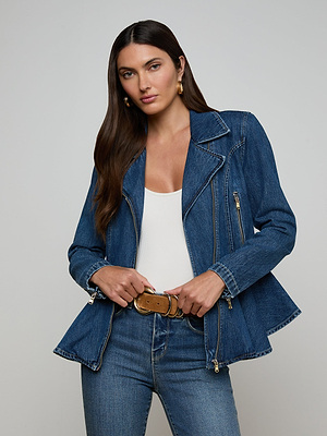A woman with long brown hair wears the Moxie Peplum Biker Jacket over a white top, paired with blue jeans, gold hoop earrings, and a tan belt. She stands against a plain light background, looking confidently at the camera.