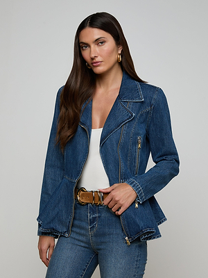 A woman with long brown hair wears the Moxie Peplum Biker Jacket over a white top and blue jeans, accessorized with gold hoop earrings and a large gold-buckle belt, standing against a plain light gray background.