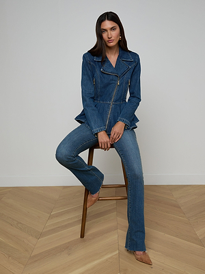 A woman with long dark hair sits on a wooden stool against a plain wall, wearing the Moxie Peplum Biker Jacket in blue denim and matching flared jeans with tan pointed-toe heels on a light wood floor.