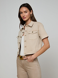 A woman wearing the Kirstie Jean Shirt Jacket in beige, styled over a white top and matching beige pants, poses with one hand in her pocket against a plain light background. This cropped denim jacket adds a modern touch to her outfit.