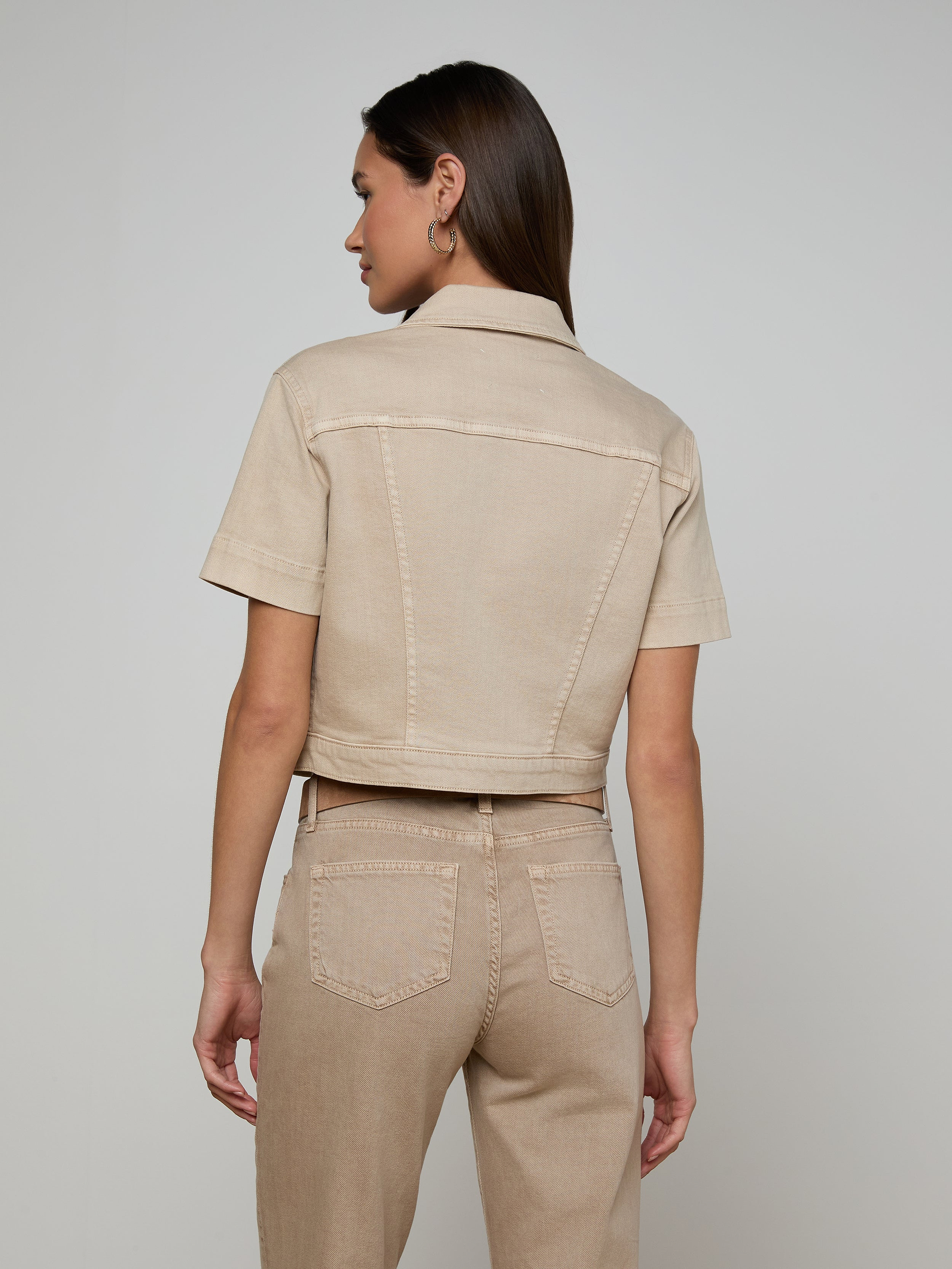 A woman stands facing away, wearing the beige Kirstie Jean Shirt Jacket with matching high-waisted pants. The background is plain and light-colored.