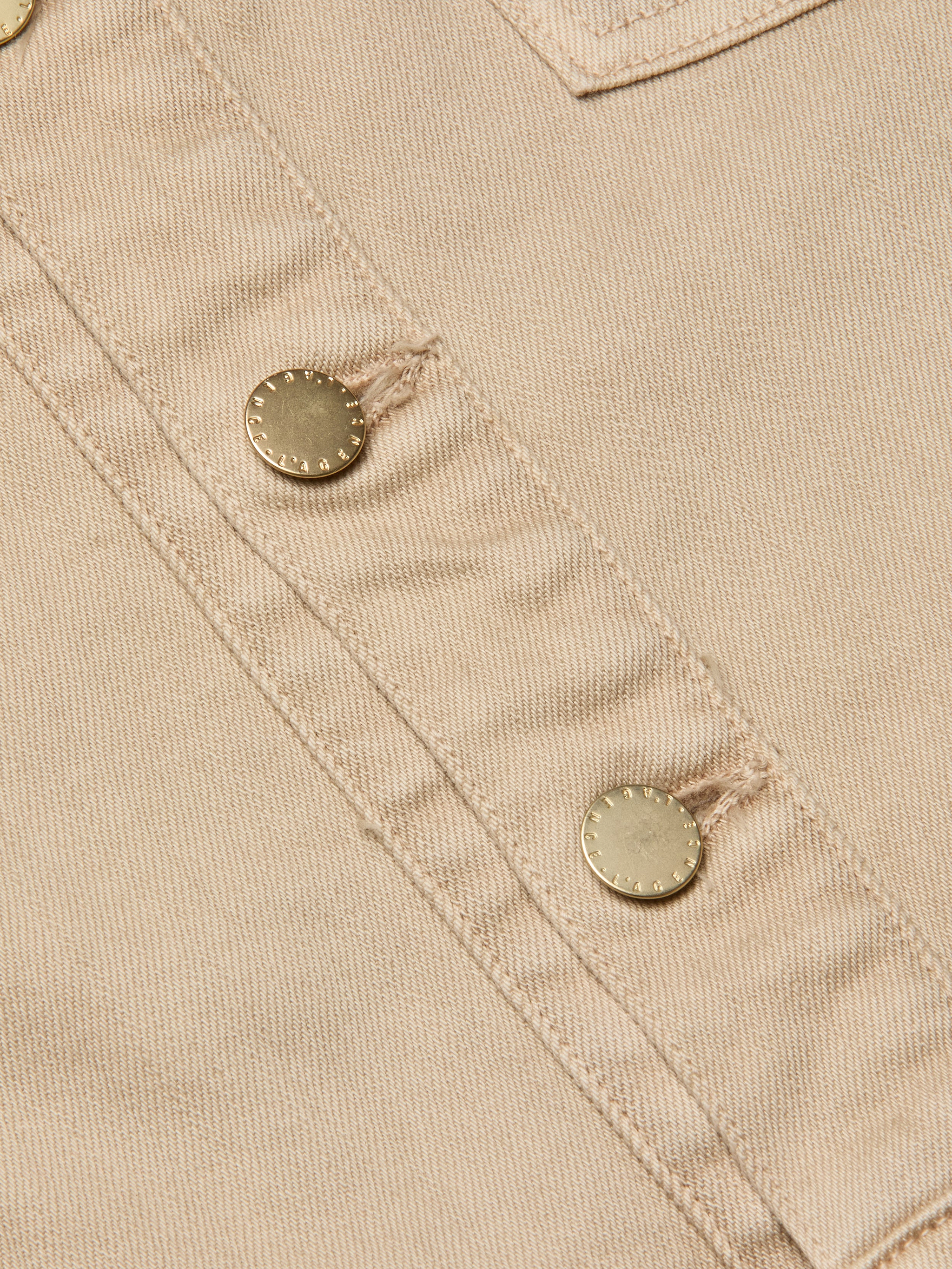 Close-up of beige fabric featuring two metallic buttons sewn in a diagonal line, highlighting the visible stitching and textured detailing characteristic of the Kirstie Jean Shirt Jacket.